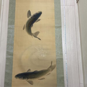 以下が含まれることがあります： クリーム色の背景に、グレーと黒の鯉が2匹描かれた伝統的な日本の巻物。巻物には緑色の縁と金色の装飾が施されています。