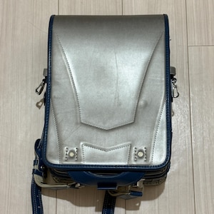 Puede incluir: Una mochila escolar estructurada con una solapa plateada metálica y ribete azul oscuro. El bolso presenta una silueta rectangular, un asa superior y dos tachuelas metálicas en la parte inferior de la solapa. Incluye correas ajustables azul oscuro.