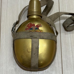 May include: A gold-coloured metal canteen with a matching screw-top cap and a beige fabric strap harness. A small, weathered sticker featuring red and blue graphic elements and Japanese text is adhered to the upper centre of the canteen body.