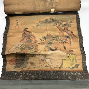 May include: Antique Asian scroll painting depicting figures in a landscape. The artwork features a muted color palette with figures in various poses, a tree, and a fish. The scroll is rolled up at the top and has a decorative border.