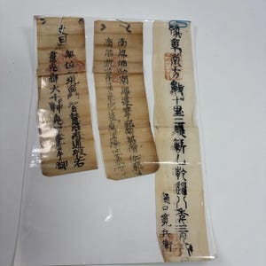 May include: Three antique, handwritten scrolls with black ink calligraphy, protected in a clear plastic sleeve. The aged paper has a light brown hue. The text is in an Asian language, with red seals and stamps visible.