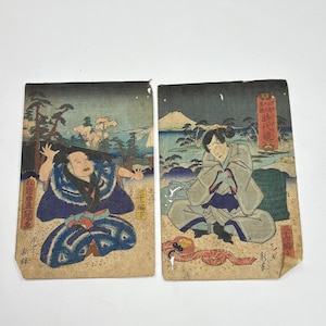 以下が含まれることがあります： 2つのアンティーク日本の木版画。左の版画は、風景を背景にした青い着物を着た男性を描いています。右の版画は、山と水の背景を持つ着物を着た人物を示しています。両方の版画には日本語のテキストがあります。