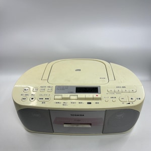May include: A cream-coloured Toshiba portable stereo system featuring a top-loading CD player, a cassette deck, and an AM/FM radio. The device includes a digital display, integrated speakers, and various control buttons with Japanese text labels.
