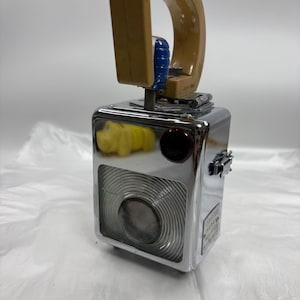 Puede incluir: Una linterna rectangular vintage de metal cromado con un asa de plástico color canela, una lente de cristal circular con anillos concéntricos, un botón circular rojo oscuro y una pequeña figura de animal amarilla visible.