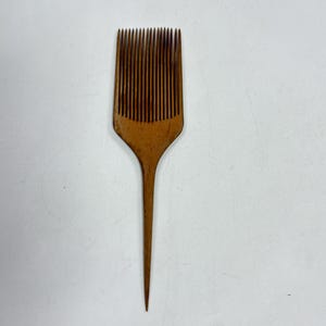 May include: A wooden hair pick featuring a wide, rectangular head with numerous thin, closely spaced teeth and a long, pointed handle. The natural wood grain is visible throughout the surface, displaying a warm, medium-brown color tone.