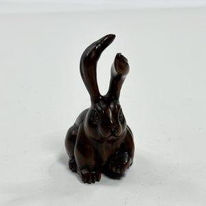 May include: A detailed, dark brown wooden rabbit figurine with upright ears. The rabbit is carved with visible fur texture and sits in a relaxed pose. The figurine is set against a plain white background.