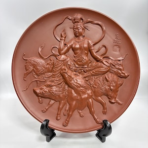 Puede incluir: Un plato de cerámica color terracota con una escultura en relieve detallada. La obra de arte representa una figura sentada sobre tres jabalíes, con intrincados detalles en la ropa y los elementos circundantes. El plato se exhibe sobre un soporte negro.