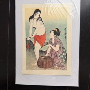 以下が含まれることがあります： 浮世絵スタイルのイラストが描かれたマット付きアートプリント。水辺にいる二人の人物が描かれています。一人は長い黒髪で口に刃物をくわえ、赤い布を巻いて立っています。もう一人は紫色の柄の着物を着て膝をつき、編み込まれた籠のそばで貝殻を持っています。
