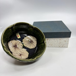 May include: A ceramic bowl with a dark green exterior and a dark blue interior. The bowl features a floral design with three beige and brown lily pads. A matching box is in the background.