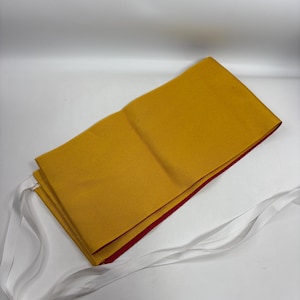 May include: A rectangular mustard yellow fabric apron featuring a contrasting red inner lining and long white fabric ties attached to the sides. The apron is folded neatly, showcasing the vibrant yellow exterior and the thin red edge along the bottom.