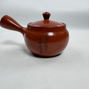 May include: A reddish-brown ceramic teapot with a side handle and a matching lid. The body of the teapot features black calligraphic text written vertically on the front surface. The design includes a rounded base and a small knob on the lid.
