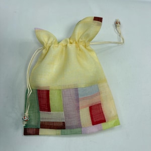 May include: A small, rectangular drawstring pouch made of sheer, light yellow fabric. The lower section features a patchwork design of rectangular panels in shades of red, green, purple, and light blue. It includes thin white drawstrings with knots.