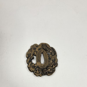May include: A bronze-colored Japanese katana tsuba, a sword guard, with a dragon motif. The tsuba features a central opening for the sword tang and is surrounded by a detailed relief of a dragon amidst swirling clouds. The overall design is ornate and detailed.