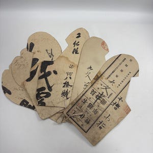 以下が含まれることがあります： 濃い茶色の日本語の書道と印刷されたテキストが施された、ベージュ色の扇形の紙タグのコレクション。タグは重ねられ、底で細い紐で結ばれており、経年変化による薄茶色のシミや摩耗が見られます。