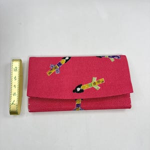 May include: A rectangular pink fabric wallet featuring a repeating pattern of stylized figures in traditional attire. To the left, a gold-colored rectangular tag is visible with Japanese text printed vertically in black ink.