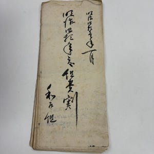 May include: A rectangular stack of aged, off-white paper sheets featuring vertical columns of black ink calligraphy. The paper edges appear worn and slightly frayed, with visible signs of discoloration and age spots across the surface.