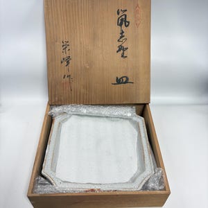 以下が含まれることがあります： ユニークな不規則なエッジを持つ正方形のオフホワイトのセラミックプレートが、開いた木箱の中にあります。プレートは保護プラスチックで包まれています。木箱の蓋には日本の書道があります。