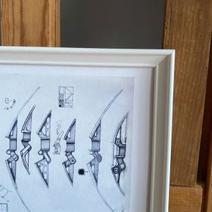 May include: A framed monochrome architectural drawing of several bow designs. The artwork, in a white frame, presents detailed sketches of different bow styles, including their components and measurements in cm. The drawing is in black and white.