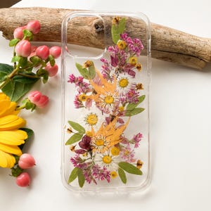 May include: Clear phone case with pressed flowers. The case features a vertical arrangement of white, yellow, and purple flowers, with green leaves and orange accents. The case is made of clear plastic.