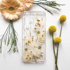 May include: A clear phone case with dried flowers and foliage encased within. The case features white and yellow flowers, with brown and green stems. The background is white, with additional flowers and greenery around the case.