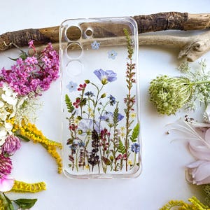 Op de afbeelding: Transparante telefoonhoes met geperste bloemen in blauwe, paarse en witte tinten. De hoes heeft een rechthoekige vorm met uitsparingen voor de camera en knoppen. De bloemarrangement is natuurlijk en biedt een uniek, artistiek ontwerp.