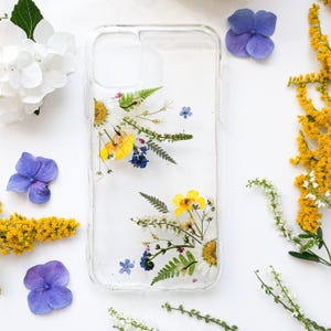 May include: Clear phone case with embedded dried flowers, including yellow, white, and blue blossoms. The case is surrounded by loose flowers on a white surface, creating a floral theme.