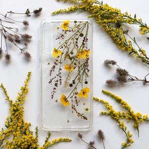 Può includere: Custodia trasparente per telefono con fiori gialli e verde incorporati. La custodia ha una forma rettangolare con angoli arrotondati. Lo sfondo è bianco, con ulteriori rametti floreali gialli disposti attorno alla custodia.