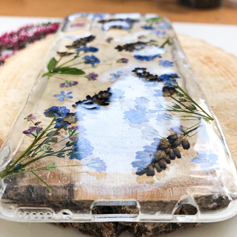 May include: Clear phone case with embedded dried flowers. The case features blue and purple flowers, and brown seed heads, set against a light-colored background. The case is made of clear plastic and is designed to protect a phone.