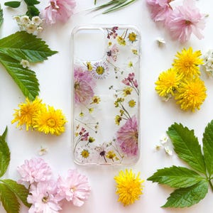 Könnte beinhalten: Transparente Handyhülle mit gepressten Blumen, darunter rosa, gelbe und weiße Blüten. Die Hülle ist von frischen Blumen und grünen Blättern auf weißem Grund umgeben. Ein florales Design.