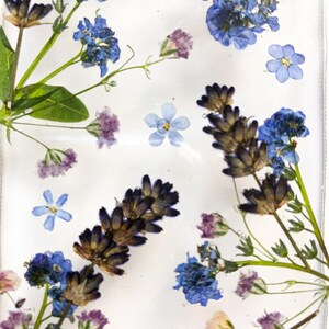 May include: A clear, rectangular object filled with dried flowers. The arrangement includes blue forget-me-nots, lavender, and small purple flowers. Green stems and leaves are also visible, set against a white background.