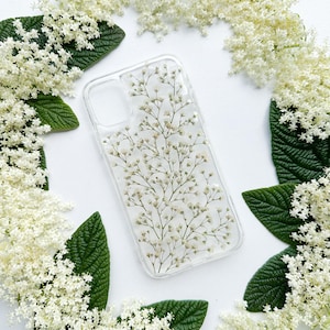 May include: Clear phone case with delicate white flowers and green stems encased within. The case is surrounded by white flowers and green leaves, creating a natural, floral theme.