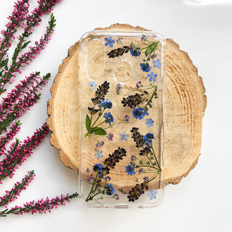 May include: Clear phone case with embedded dried flowers. The case features blue and purple flowers and green leaves. The case is displayed on a wooden slice, with pink and green sprigs in the background.