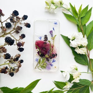 Può includere: Una custodia trasparente per telefono con fiori e vegetazione pressati. La custodia presenta un grande fiore rosso intenso, piccoli fiori blu e viola e steli verdi. More e fiori bianchi sono disposti attorno alla custodia su uno sfondo bianco.