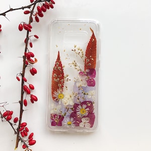 May include: Clear phone case with pressed flowers and gold flakes. The case features a variety of dried flowers in shades of purple, white, and yellow, with two large orange leaves. The case is rectangular and fits a smartphone.