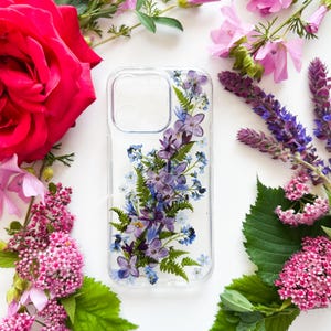 May include: Clear phone case with a floral design featuring purple, blue, and green flowers and foliage. The case is surrounded by pink and purple flowers and green leaves on a white background.