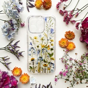 Op de afbeelding: Een doorzichtig smartphonehoesje met een ingebed arrangement van geperste wilde bloemen, waaronder witte madeliefjes, kleine blauwe bloesems en gele blaadjes, met een vierkante uitsparing voor de camera in de linkerbovenhoek.