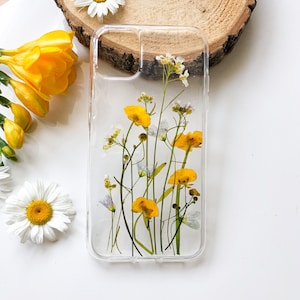 Puede incluir: Funda transparente para teléfono con flores silvestres amarillas y blancas prensadas. La funda presenta un diseño transparente, mostrando la belleza natural de las flores. Flores amarillas y margaritas rodean la funda.