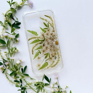 Könnte beinhalten: Transparente Handyhülle mit gepressten Blumen, darunter eine Gänseblümchen, Farne und andere getrocknete Pflanzen. Die Hülle ist rechteckig mit abgerundeten Ecken und einem Kameraausschnitt. Frische Blumen umgeben die Hülle auf weißem Hintergrund.