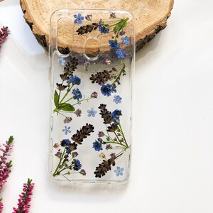 May include: Clear phone case with embedded dried lavender and blue forget-me-nots. The flowers are arranged in a natural, organic pattern. The case is rectangular with cutouts for the camera and other phone features.
