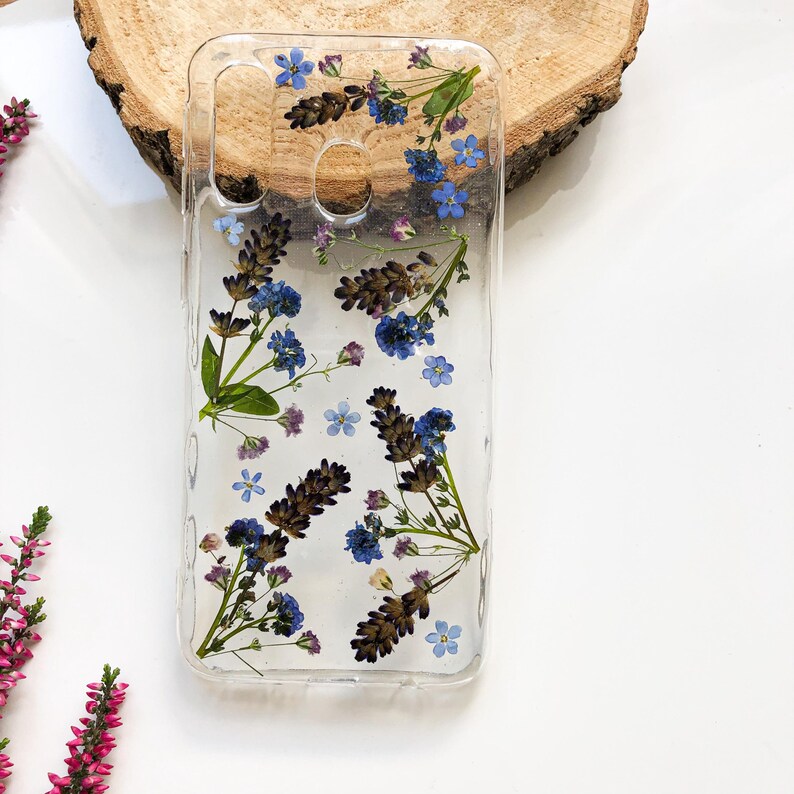 May include: Clear phone case with embedded dried lavender and blue forget-me-nots. The flowers are arranged in a natural, organic pattern. The case is rectangular with cutouts for the camera and other phone features.