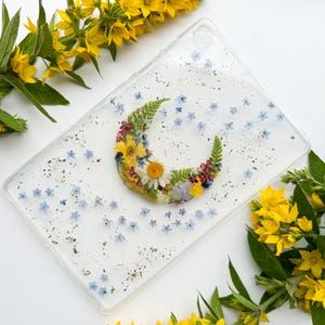 May include: Clear tablet case with a crescent moon design made of dried flowers in yellow, white, red, and blue. The case is decorated with small blue flowers and silver glitter.