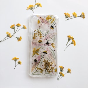 May include: Clear phone case with pressed flowers, including yellow, white, and purple blooms. The case is rectangular and has cutouts for the camera and buttons. Yellow flowers are scattered around the case on a white background.