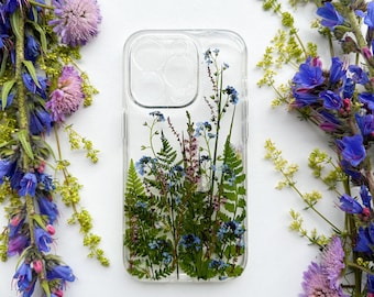 May include: A clear phone case with pressed flowers, including ferns, blue and purple wildflowers. The case is surrounded by blue and purple flowers and green foliage against a white background.
