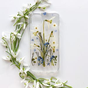 Può includere: Custodia trasparente per telefono con fiori pressati, tra cui bucaneve bianchi e piccoli fiori blu. La custodia contiene una varietà di fiori secchi, racchiusi in resina trasparente. Fiori freschi di bucaneve circondano la custodia.