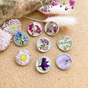 May include: A collection of eight round, silver-toned resin pendants, each encapsulating a different pressed flower. The flowers vary in color, including pink, purple, blue, and white, and are set against a clear background.