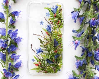 May include: Clear phone case with a floral design featuring pressed blue flowers, green ferns, and red accents. The case is surrounded by vibrant purple flowers on a white background, creating a natural aesthetic.
