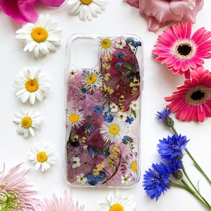 Op de afbeelding: Transparante telefoonhoes met ingebedde gedroogde bloemen, waaronder witte madeliefjes, blauwe korenbloemen en roze bloemblaadjes. De hoes is omgeven door verse bloemen, wat een bloemige en natuurlijke uitstraling creëert.