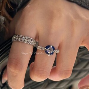 May include: Two rings are shown on a finger. One is a silver band with a repeating diamond pattern. The other is a silver ring with a large, round, dark blue stone and smaller clear stones. The text "Design By Viere Lux" is visible.