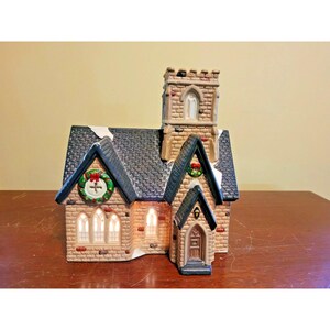 May include: A ceramic miniature church building with tan brick walls, a dark gray shingled roof with white snow accents, and a tall square tower. The structure features arched windows with warm interior lighting, a front door, and green wreaths.
