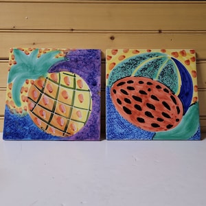Może przedstawiać: Dwa kolorowe ceramiczne kafelki z wzorem ananasa i czerwonego owocu. Kafelka z ananasem ma żółty i zielony ananas na niebieskim tle. Kafelka z czerwonym owocem ma czerwony owoc z czarnymi pestkami na zielono-niebieskim tle.